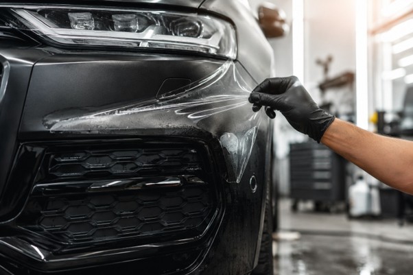 A close-up of a matte black SUV's front bumper with a technician's gloved hand applying clear paint protection film, sunlight reflecting off the semi-applied film edge, professional automotive detailing studio background, shallow depth of field, photorealistic.