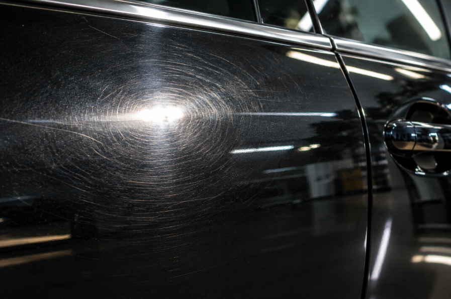 Close-up of a car door panel showing fine web-like swirl marks catching direct light, glossy black or dark paint surface, professional automotive detail photography style