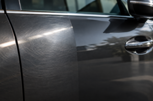 Close-up of a car door panel showing faint swirl marks catching light on one side, smooth glossy finish on the other, soft directional lighting, automotive detail photography style